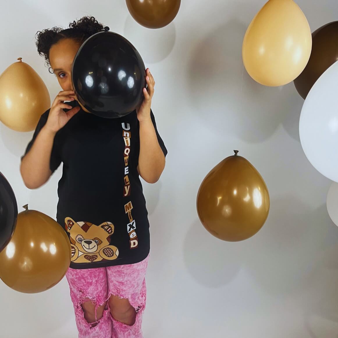 Child wearing Uniquely Mixed black t-shirt with vertical letters 'Uniquely Mixed' and teddy bear graphic, paired with pink ripped jeans surrounded by brown, white, and black balloons, representing inclusive kids fashion and playful streetwear style.