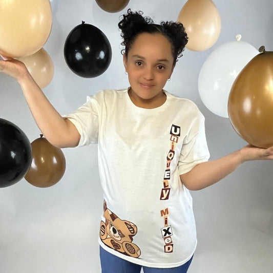 Smiling child wearing Uniquely Mixed white t-shirt with vertical logo and teddy bear graphic, surrounded by black, brown, and white balloons, representing inclusive kids fashion and playful streetwear style.