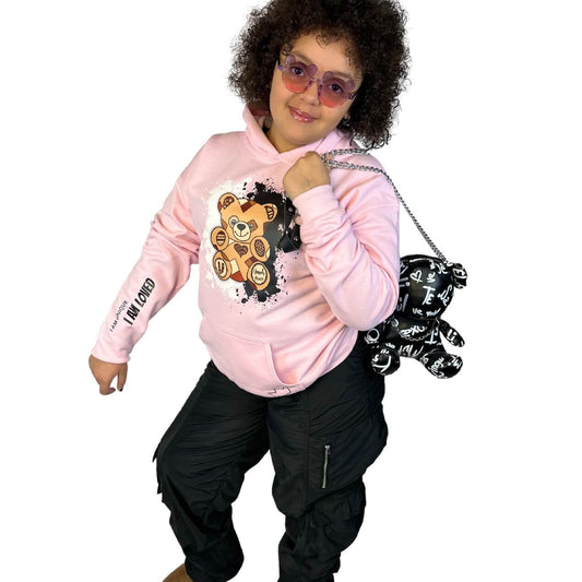 Stylish young biracial girl wearing a pink "I Am Loved" Unity Bear hoodie with a teddy bear graphic, black cargo pants, and brown boots, holding a black teddy bear purse. Confident kids' streetwear fashion promoting self-love and diversity. 